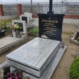 Granitowy nagrobek z czarną tablicą inskrypcyjną i krzyżem, z datami urodzin i śmierci członków rodziny Żymańczyk, otoczony innymi nagrobkami i zielenią na cmentarzu.