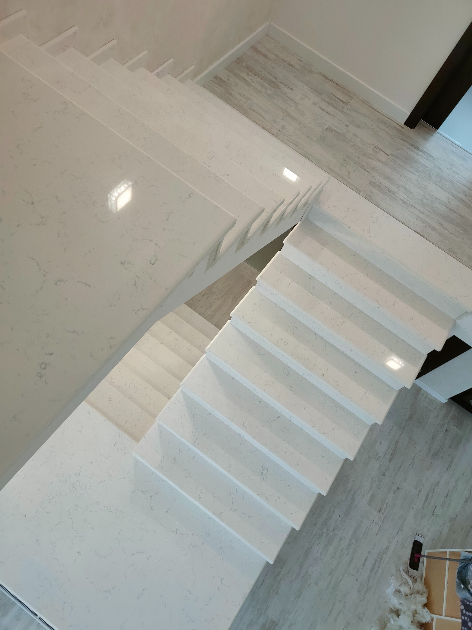 Widok z góry na jasne, eleganckie schody z materiału imitującego kamień, z subtelnym wzorem żyłek, prowadzące do jasnego pomieszczenia z podłogą w odcieniach szarości.