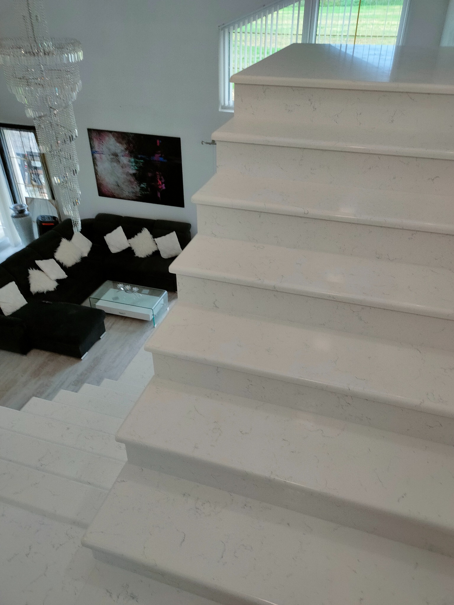 Nowoczesne, jasne schody prowadzące do salonu z ciemną sofą i kryształowym żyrandolem.