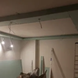 Sufit podwieszany z płyt gipsowo-kartonowych w trakcie montażu, widoczne kable oświetleniowe, drabina i płyty gotowe do użycia.