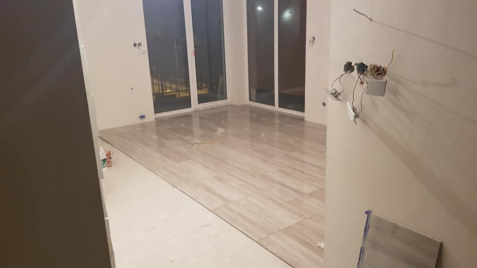 Układanie płytek podłogowych w jasnym, nowoczesnym wnętrzu z dużymi oknami balkonowymi; widoczne niedokończone prace elektryczne na ścianie.