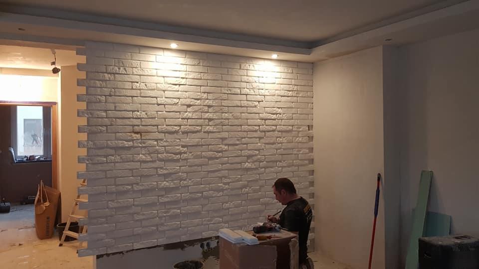 Montaż dekoracyjnej ściany z białej cegły w trakcie remontu mieszkania, widoczne narzędzia i pracownik w trakcie wykańczania krawędzi.