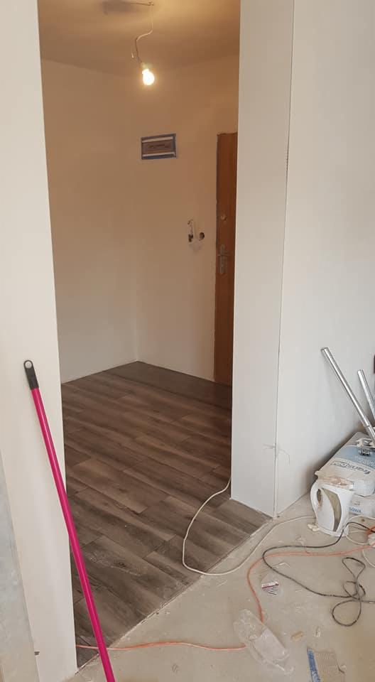 Wnętrze mieszkania w trakcie remontu: świeżo położona podłoga imitująca drewno, widoczne narzędzia i materiały budowlane, surowy stan ścian.