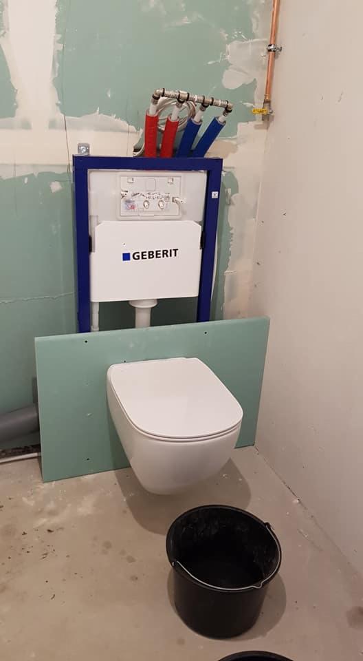 Wisząca miska WC marki Geberit w trakcie montażu, widoczne rury doprowadzające wodę z niebieską i czerwoną izolacją, surowe ściany w trakcie wykończenia, na podłodze wiadro.