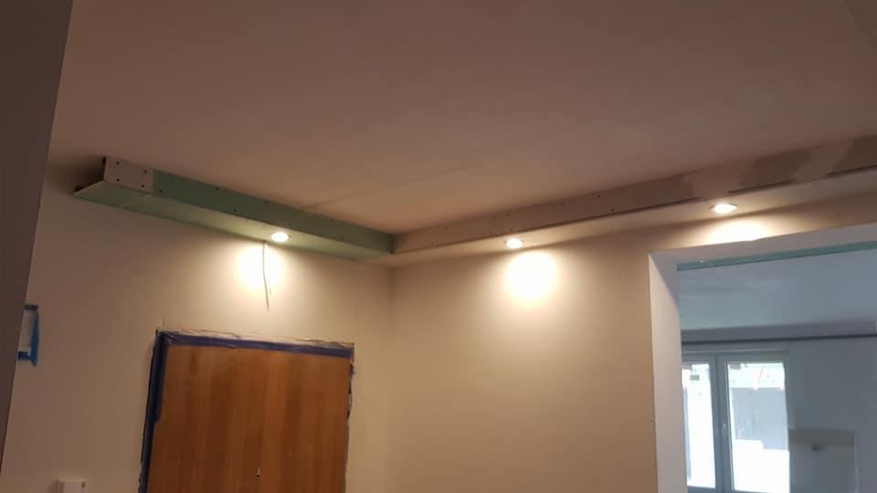 Wnętrze w trakcie prac wykończeniowych: zabudowa sufitu podwieszanego z oświetleniem punktowym, widoczne profile i płyty gipsowo-kartonowe, zabezpieczone drzwi taśmą malarską.