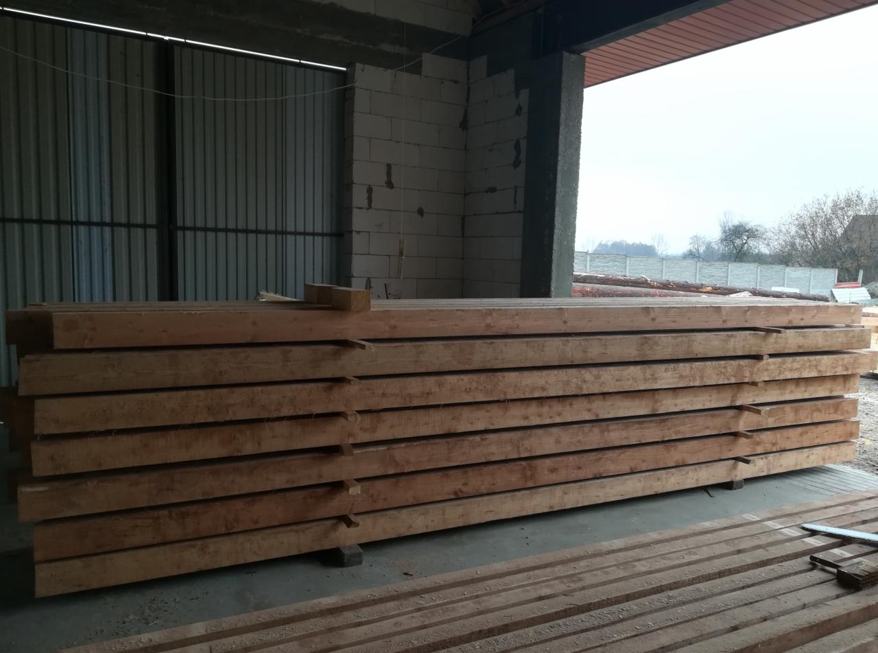 Stos drewnianych belek na placu budowy, widoczne bloczki betonowe i metalowa konstrukcja w tle.