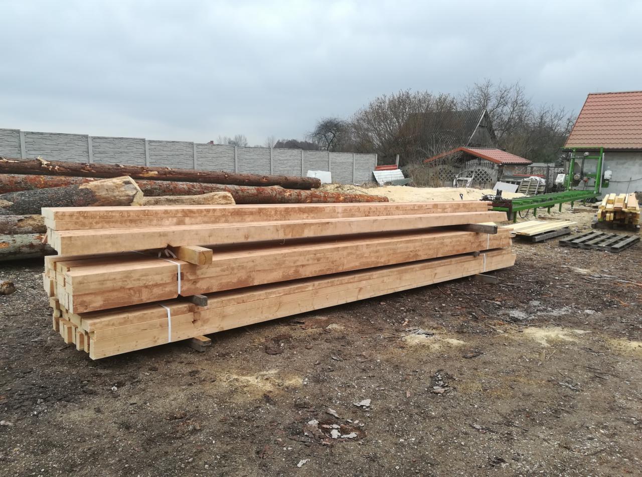 Stos świeżo przetartego drewna budowlanego na placu, w tle stosy kłód i piła taśmowa do drewna