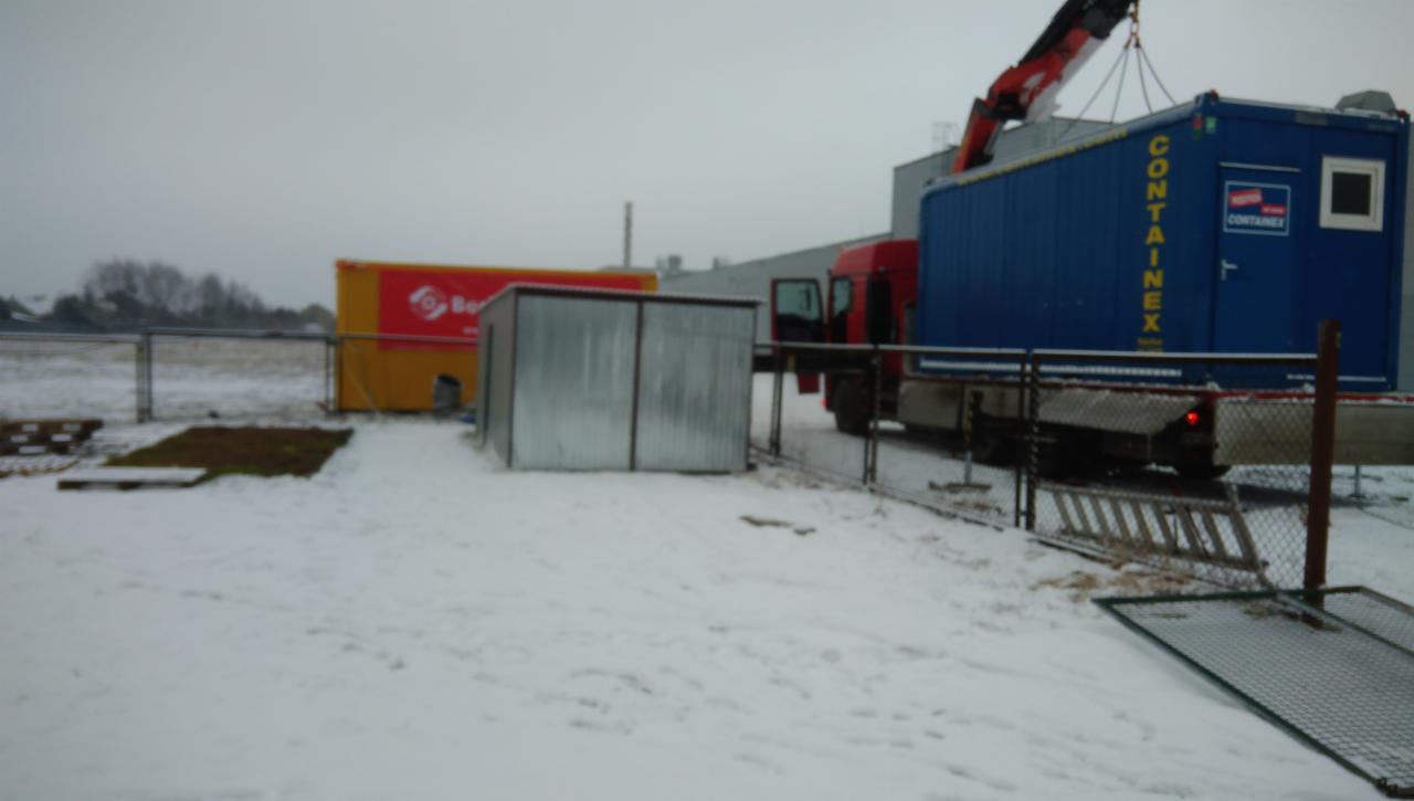 Ciężarówka z dźwigiem podnosi niebieski kontener biurowy na tle zaśnieżonego terenu i żółtego kontenera.