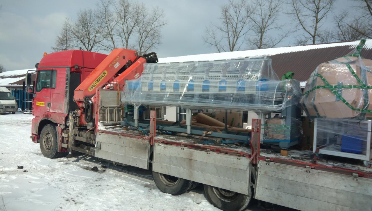 Czerwona ciężarówka z dźwigiem Palfinger przewozi zapakowane w folię urządzenie przemysłowe na platformie w zimowej scenerii.