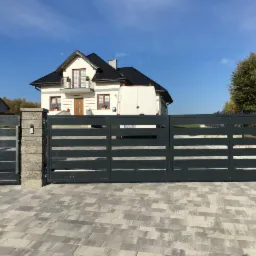 Nowoczesna, przesuwna brama wjazdowa z szarymi, poziomymi panelami na tle domu z białymi ścianami i ciemnym dachem, widok z zewnątrz, kostka brukowa.