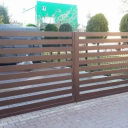 Nowoczesna brama wjazdowa z poziomymi profilami w kolorze brązowym, osadzona na słupkach z cegły klinkierowej, na tle kostki brukowej i zieleni.