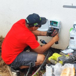 SPDE Serwis ogrodowy - Instalator w czerwonej koszulce montuje sterownik nawadniania na białej ścianie. Widoczne narzędzia, przewody i torba z nawozem. Ujęcie z boku, na zewnątrz.