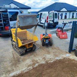 SPDE Serwis ogrodowy - Żółta mini-wywrotka z łyżką uniesioną nad ziemią, obok glebogryzarki i firmowy bus z logo na tle domu. Przygotowanie terenu pod ogród.