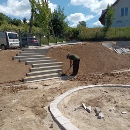 SPDE Serwis ogrodowy - Pracownik w zielonej koszulce łopatą formuje teren przy betonowych schodach zewnętrznych, wkomponowanych w ziemny nasyp. Widoczny zakrzywiony krawężnik i biały van w tle.