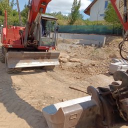 SPDE Serwis ogrodowy - Czerwona koparka kołowa z lemieszem, z łyżką w pierwszym planie, na placu budowy z częściowo wzniesionymi fundamentami z bloczków betonowych i domem w tle.