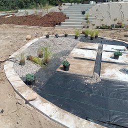 SPDE Serwis ogrodowy - Owalna rabata ogrodowa z szarym żwirem, kwadratowymi płytami i czarną agrowłókniną, w trakcie aranżacji. W tle widoczne betonowe schody i mulczowane skarpy z przygotowanymi roślinami.