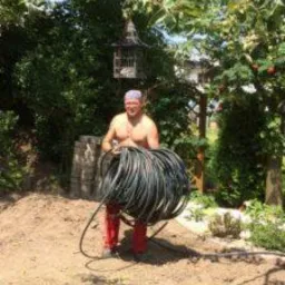 Mężczyzna bez koszulki, w czerwonych spodniach i fioletowej bandanie, trzymający zw&oacute;j czarnego węża na tle ogrodu z widoczną budką dla ptak&oacute;w i stertą betonowych kręg&oacute;w.