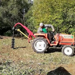 Operator na pomarańczowym traktorze z wiertnicą glebową, przygotowuje teren pod nasadzenia, widoczne drzewa w tle.