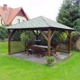 Drewniana altana ogrodowa z zielonym dachem, wyposażona w stół, ławki i grill, otoczona trawnikiem i kwiatami w donicach, z domem w tle.