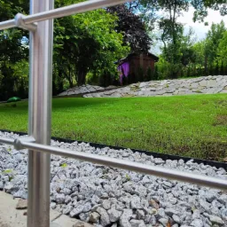 Widok na świeżo założony trawnik z rolki, ograniczony czarną obwódką i wysypany grysem, z metalową barierką w pierwszym planie oraz małym, pomalowanym na fioletowo domkiem narzędziowym w tle.