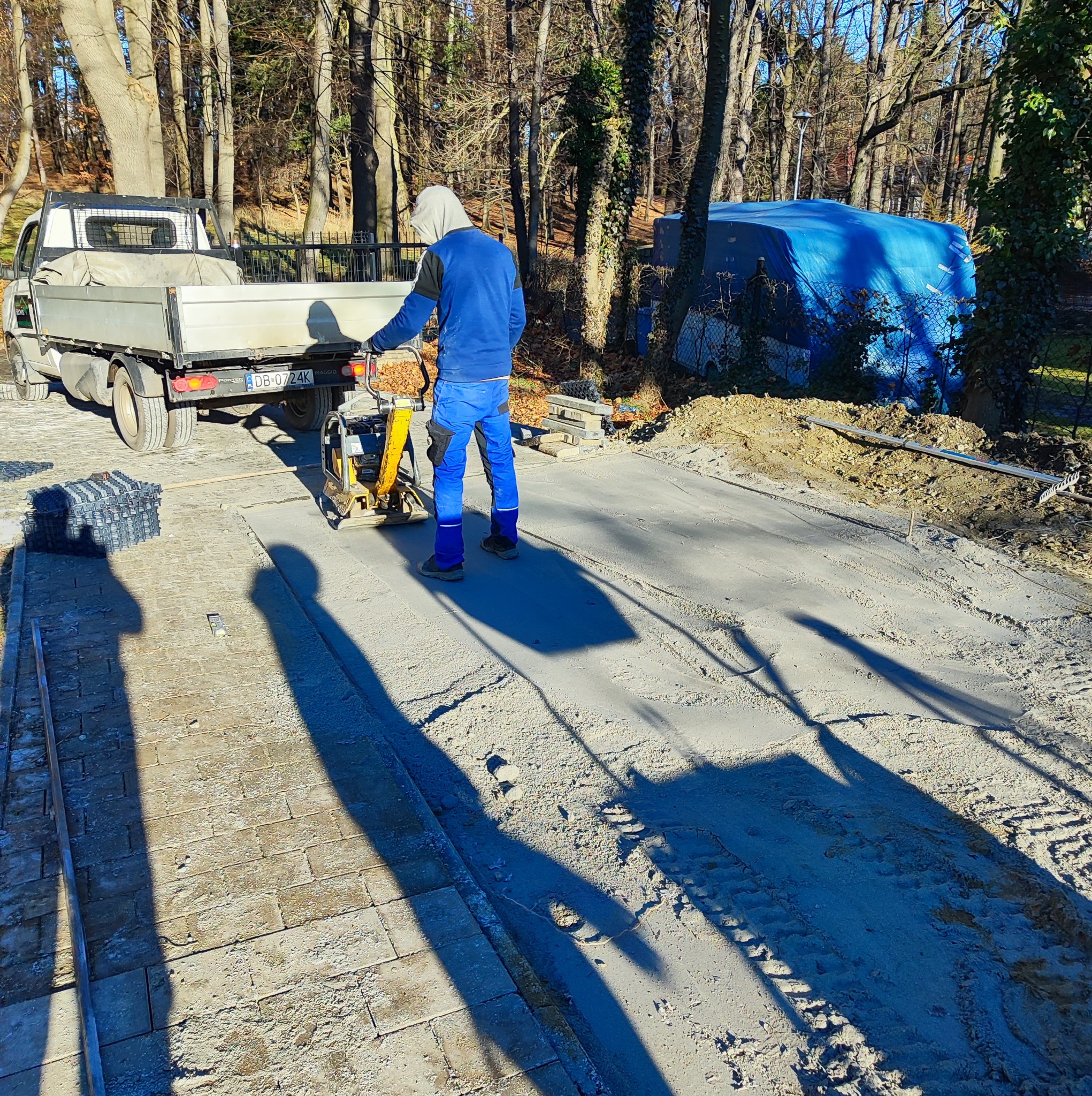 Pracownik w niebieskim kombinezonie ubija piasek zagęszczarką na przygotowanym podłożu pod kostkę brukową. W tle samochód dostawczy i drzewa.