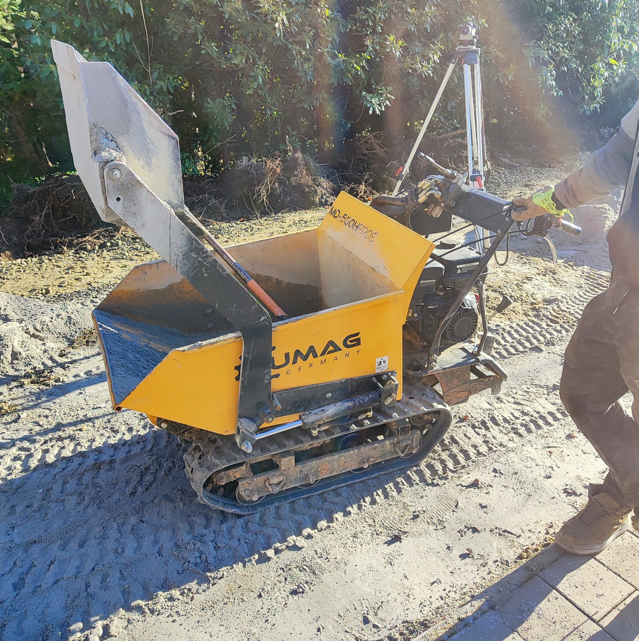 Żółta mini-wywrotka gąsienicowa UMAC Germany z podniesioną łyżką, wypełniona ziemią, na placu budowy. W tle niwelator i zieleń.