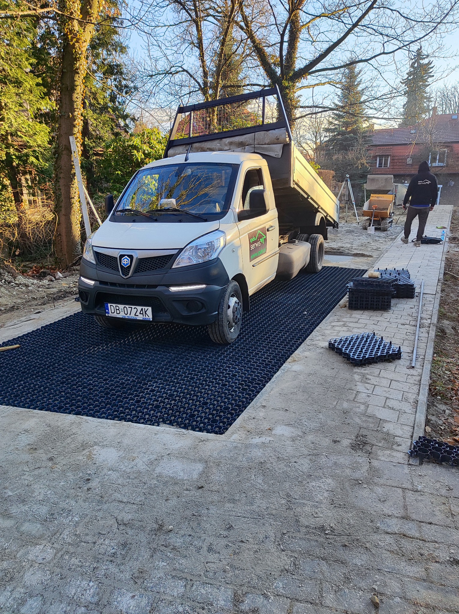 Ciężarówka na czarnych panelach drenażowych na placu budowy. W tle drzewa i budynek. Widoczna osoba w bluzie z logo firmy. Ujęcie z niskiej perspektywy.