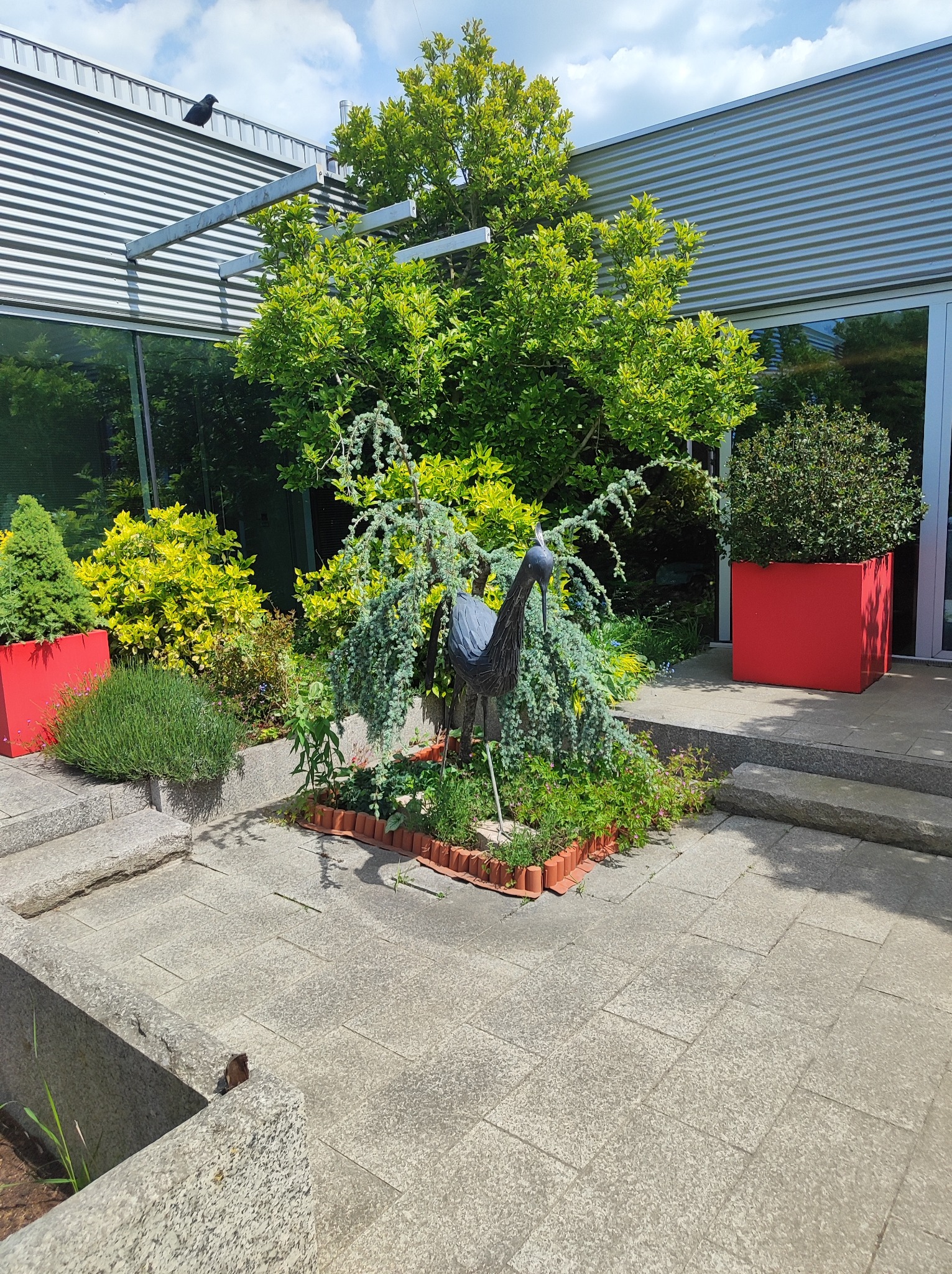 Ogród przy budynku z metalową rzeźbą czapli, roślinami w czerwonych donicach i ozdobnymi obrzeżami rabat. Nowoczesna architektura w tle, błękitne niebo.