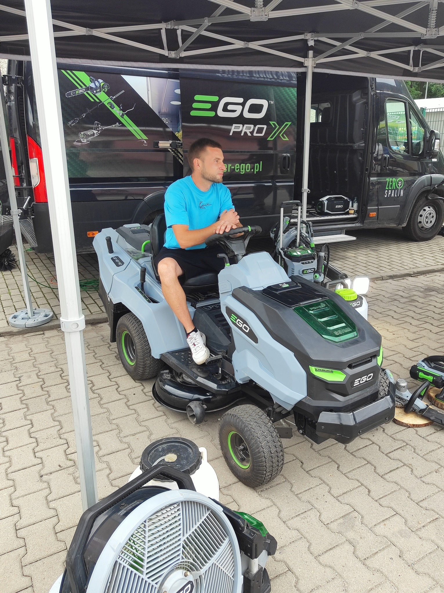 Mężczyzna siedzi na szaro-czarnej kosiarce EGO z zielonymi akcentami. W tle czarny van z logo EGO Pro X i hasłem Zero Spalin. Prezentacja sprzętu ogrodniczego.
