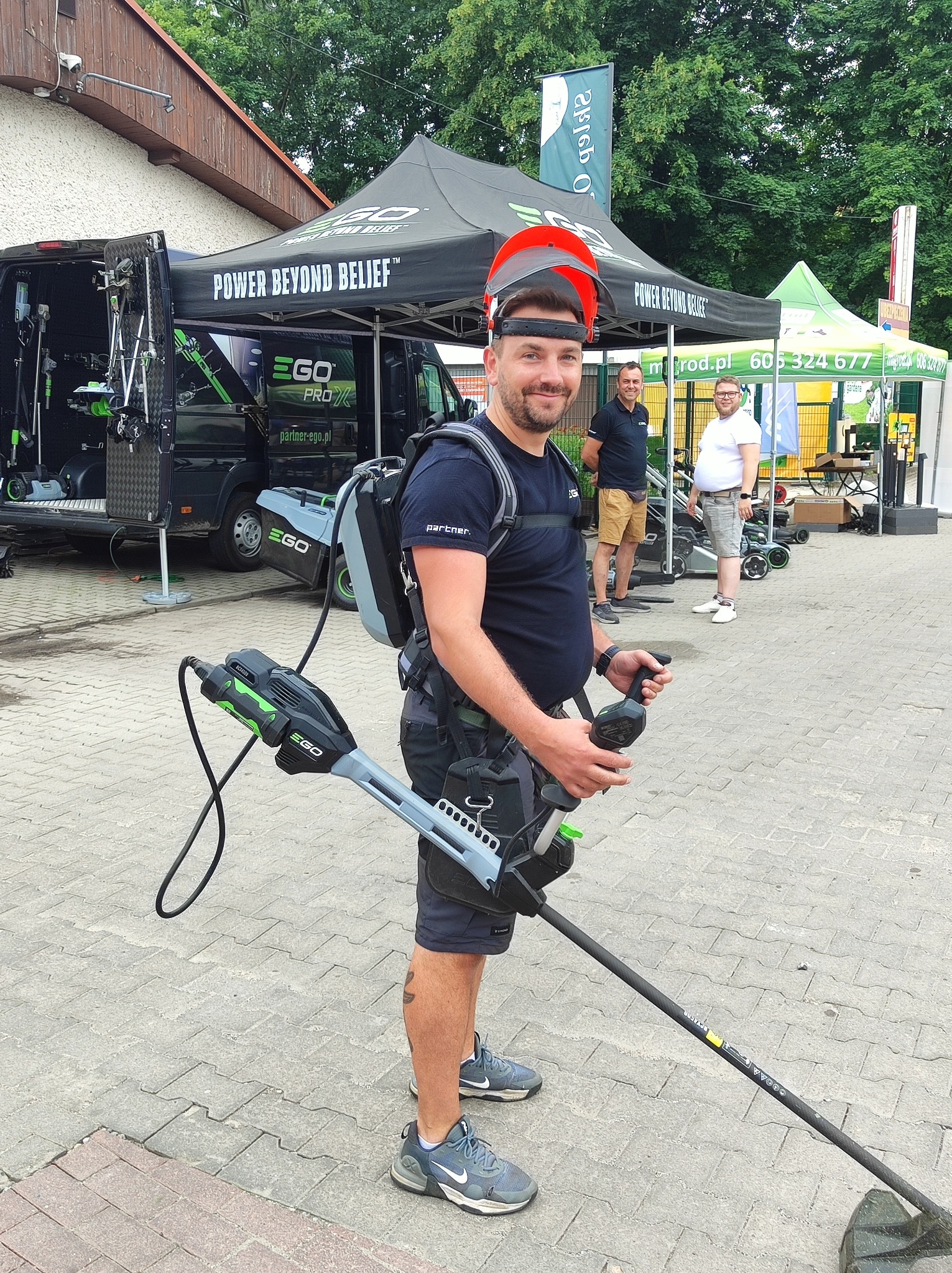 Uśmiechnięty mężczyzna w kasku z podniesioną osłoną, trzymający akumulatorową podkaszarkę EGO, z plecakiem na baterie. W tle stoisko EGO i inne osoby.