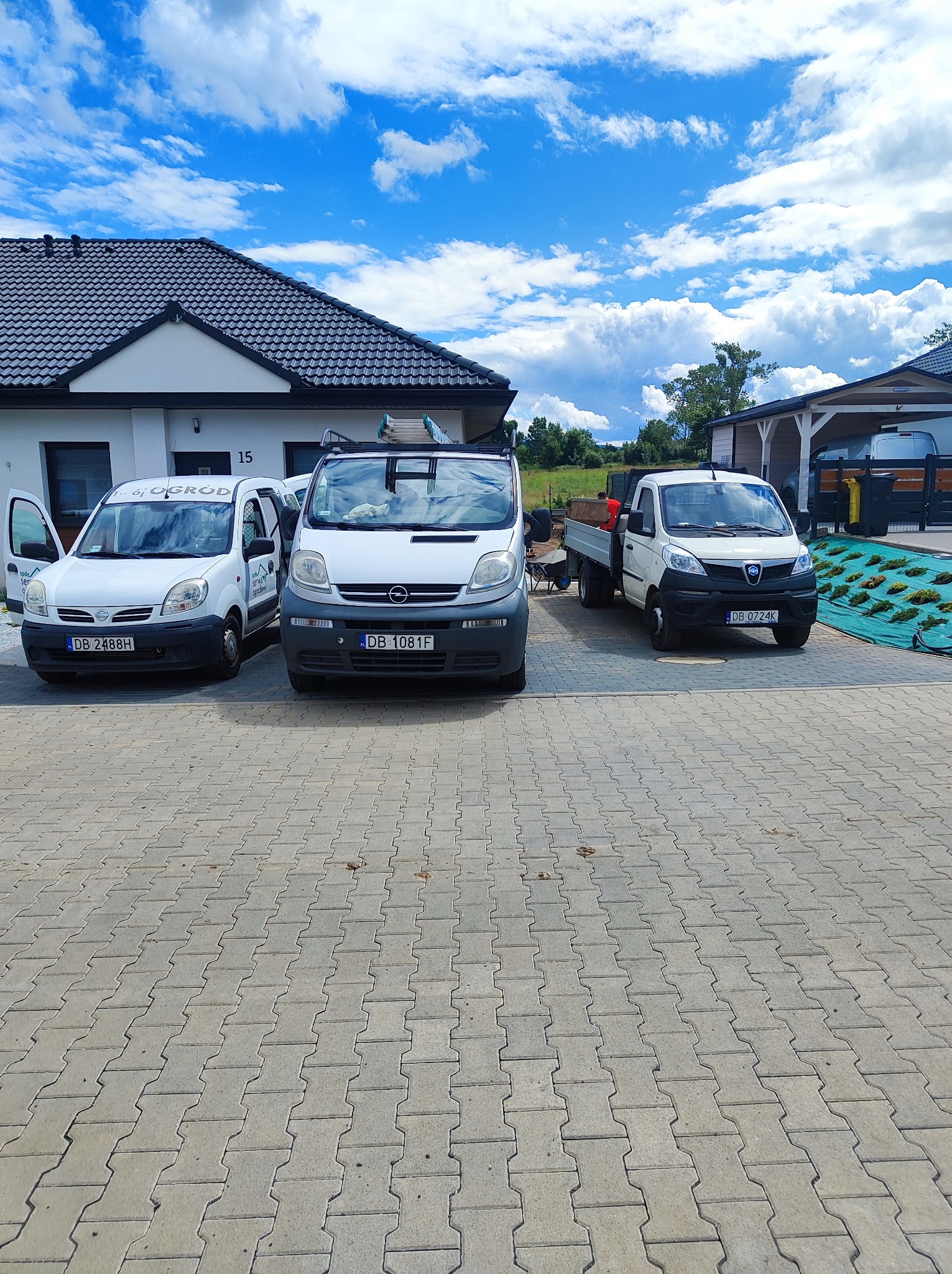 Trzy białe samochody dostawcze z logo firmy ogrodniczej zaparkowane na brukowanym podjeździe przed domem z ciemnym dachem i błękitnym niebem w tle. Jeden z samochodów ma otwarte drzwi.