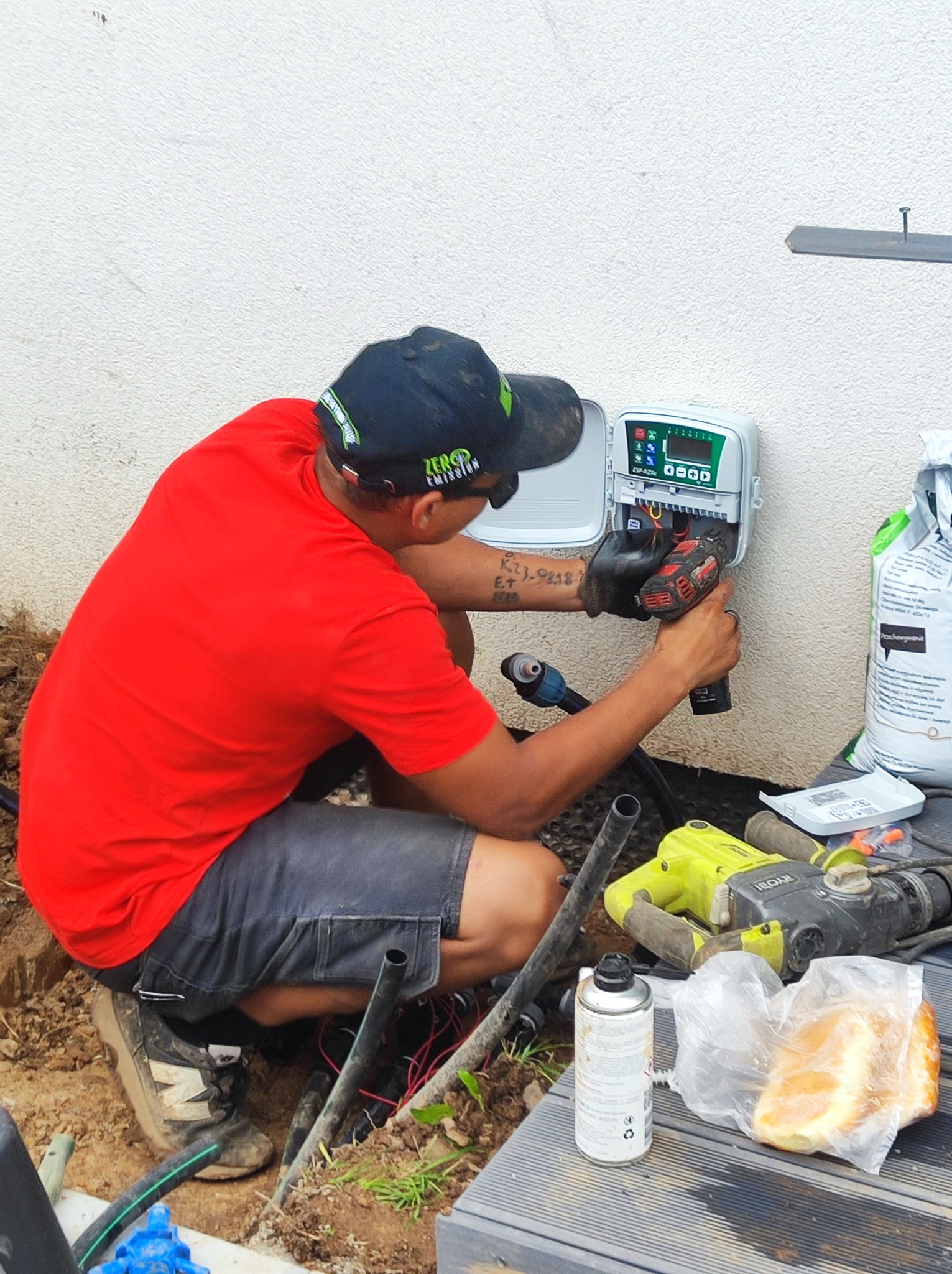 Instalator w czerwonej koszulce montuje sterownik nawadniania na białej ścianie. Widoczne narzędzia, przewody i torba z nawozem. Ujęcie z boku, na zewnątrz.