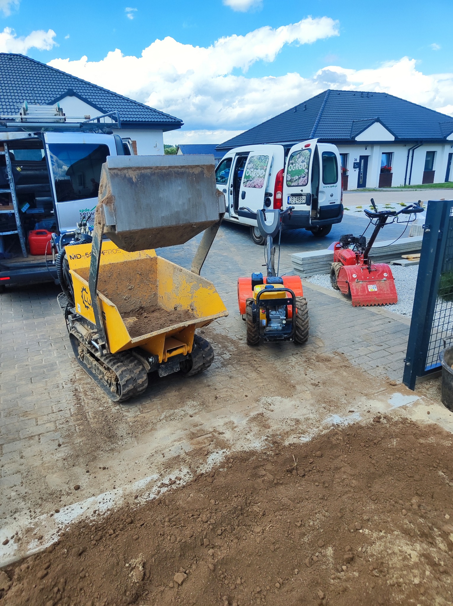 Żółta mini-wywrotka z łyżką uniesioną nad ziemią, obok glebogryzarki i firmowy bus z logo na tle domu. Przygotowanie terenu pod ogród.