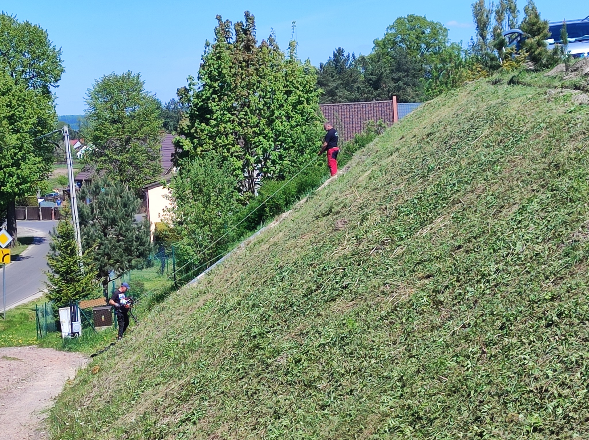 Pracownicy w trakcie koszenia stromej skarpy porośniętej trawą. Jeden z nich używa linki zabezpieczającej. W tle widoczne domy i drzewa.