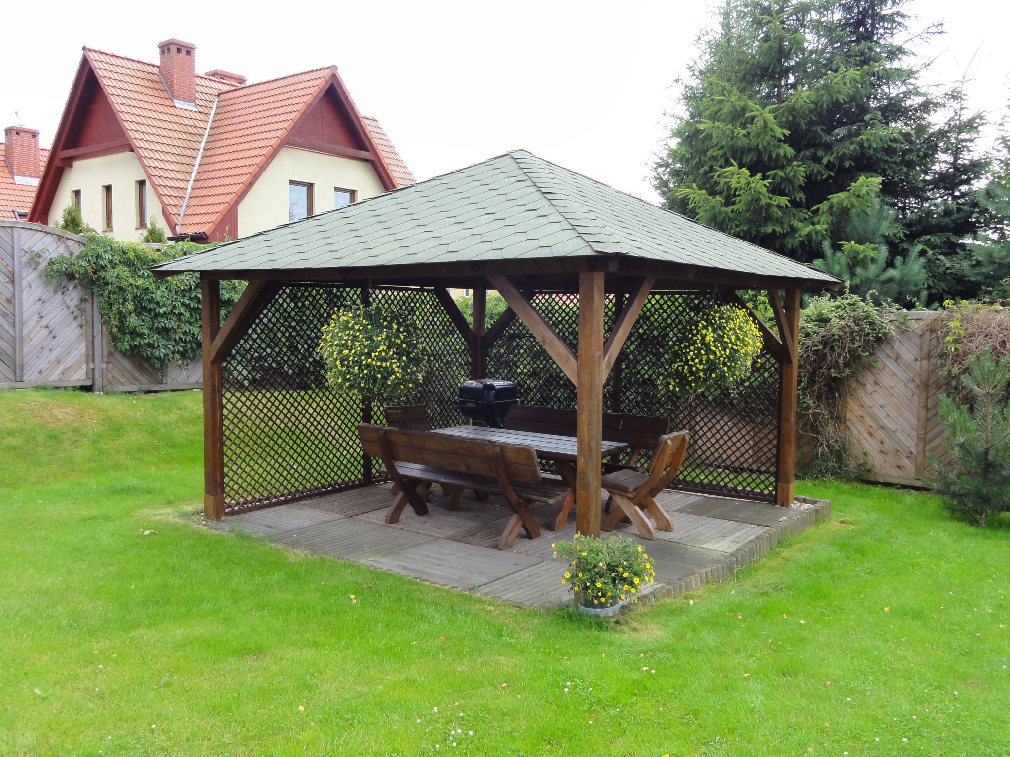 Drewniana altana ogrodowa z zielonym dachem, wyposażona w stół, ławki i grill, otoczona trawnikiem i kwiatami w donicach, z domem w tle.