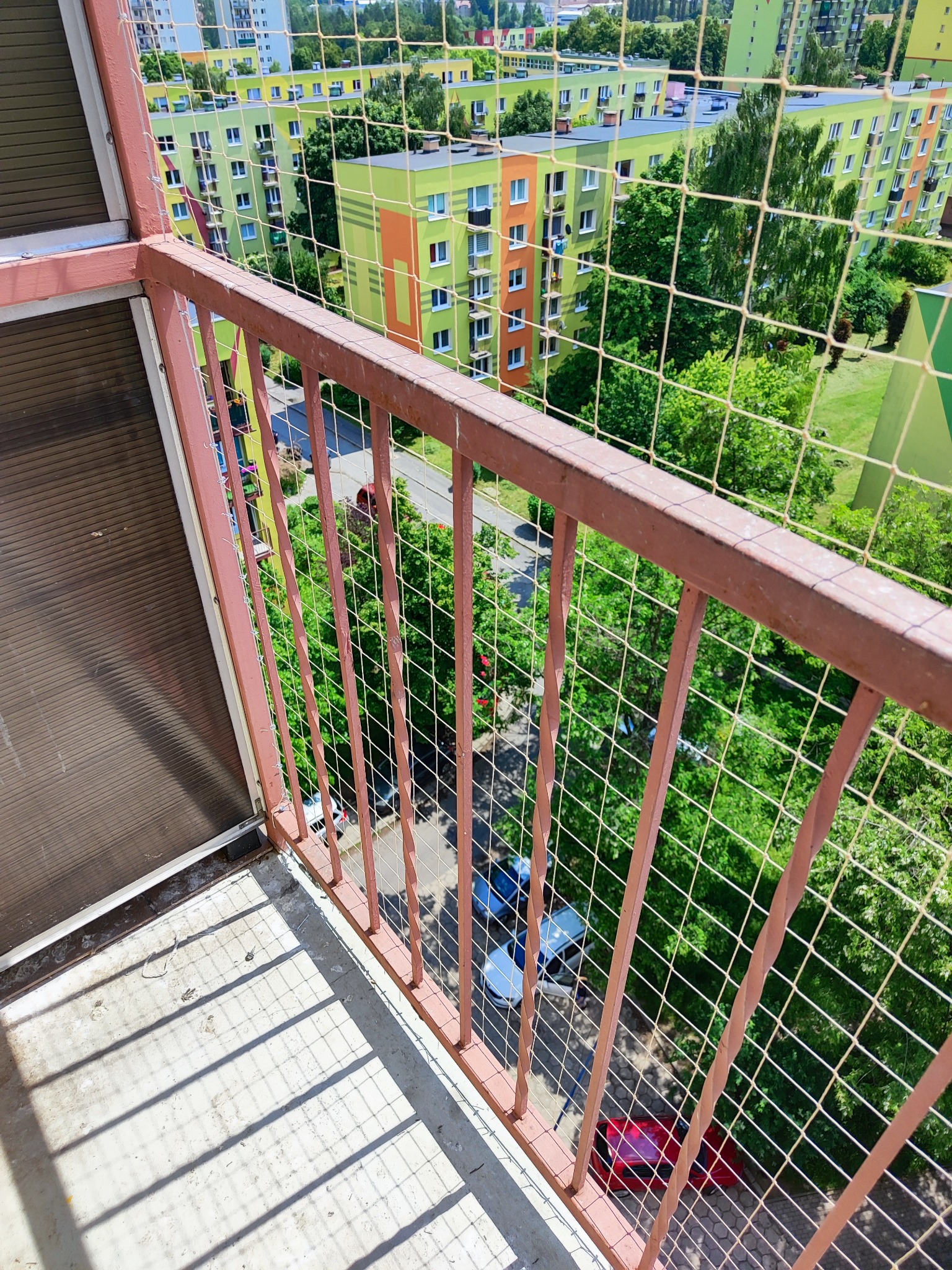 Widok z balkonu zabezpieczonego siatką ochronną na kolorowe bloki mieszkalne i zieleń drzew w słoneczny dzień.