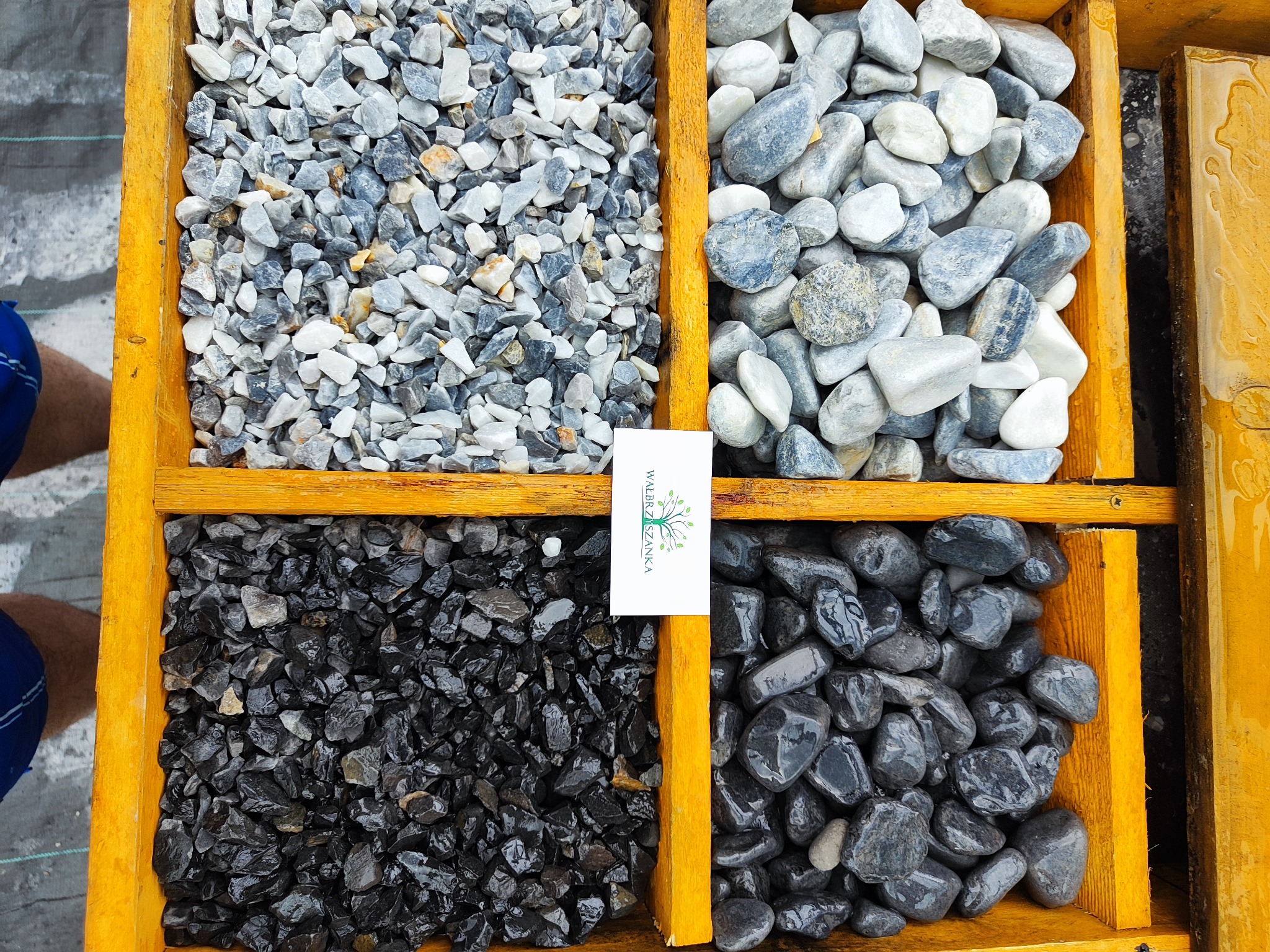 Cztery przegródki z kamieniami dekoracyjnymi: szare, biało-szare, czarne matowe i czarne błyszczące, oddzielone żółtymi listwami, z wizytówką firmy pomiędzy czarnymi kamieniami.