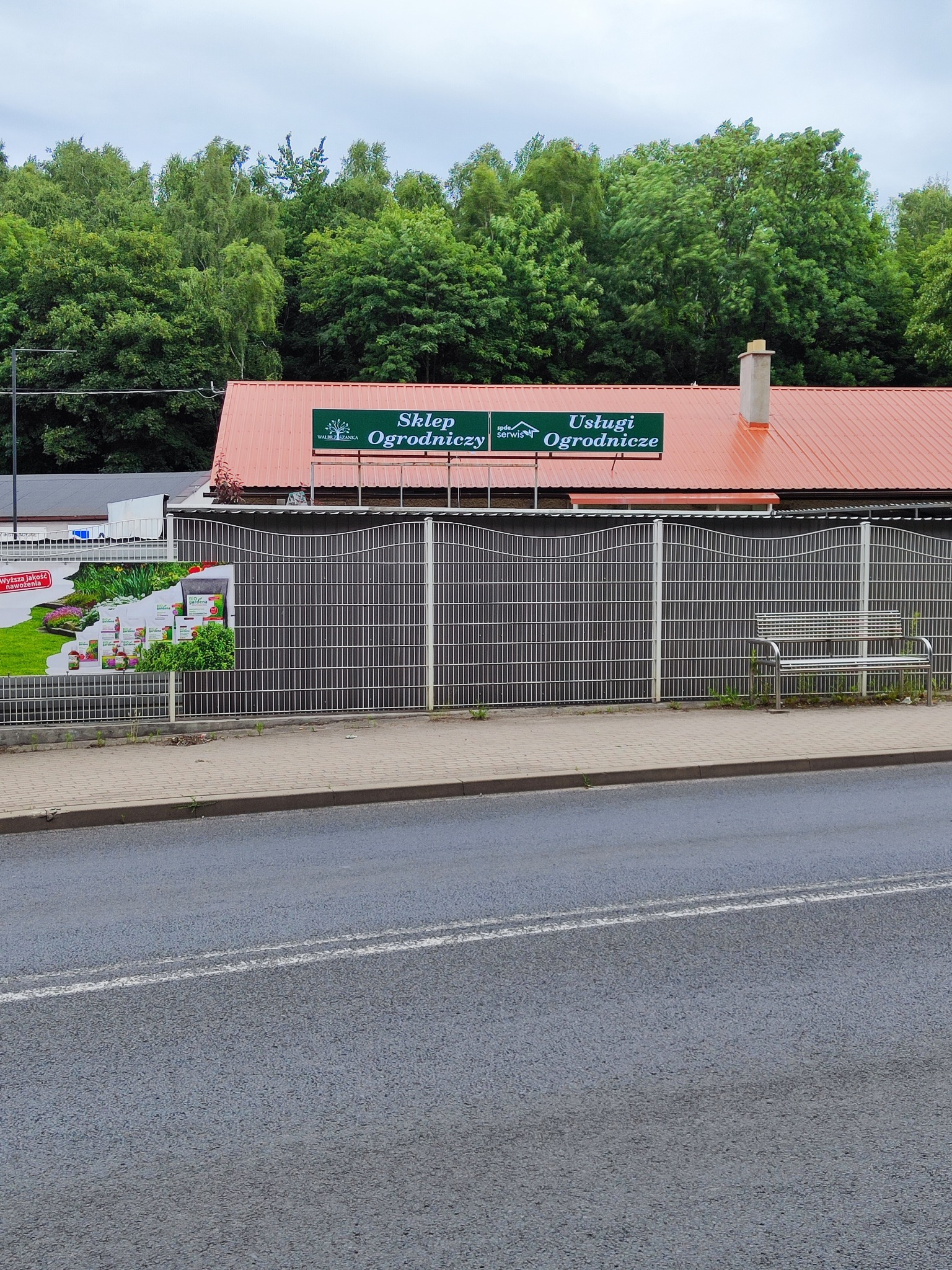 Budynek sklepu ogrodniczego z czerwonym dachem, ogrodzony metalowym płotem, na którym wisi baner reklamowy z produktami ogrodniczymi. Nad wejściem widoczny zielony szyld z napisem 'Sklep...