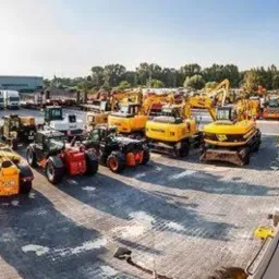 Szeroki panoramiczny widok na plac z zaparkowanymi maszynami budowlanymi, w tym żółte koparki, ładowarki, walce i ciężarówki, z budynkiem w tle i drzewami po bokach, sfotografowany w słoneczny dzień.