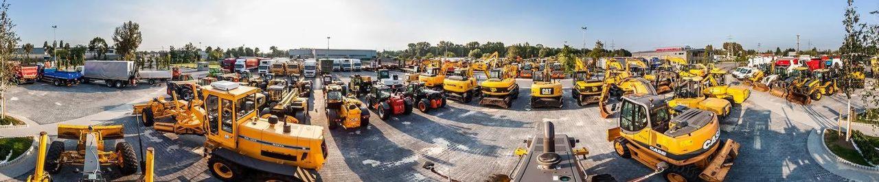 Szeroki panoramiczny widok na plac z zaparkowanymi maszynami budowlanymi, w tym żółte koparki, ładowarki, walce i ciężarówki, z budynkiem w tle i drzewami po bokach, sfotografowany w słoneczny dzień.