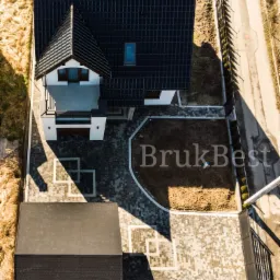 Widok z góry na dom z czarnym dachem i posesją z szarej kostki brukowej ułożonej we wzór, z widocznym logo firmy BrukBest na niezagospodarowanym terenie.