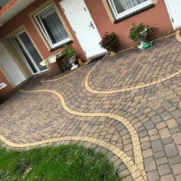 Ułożona kostka brukowa w odcieniach brązu z żółtymi obrzeżami tworzącymi falisty wzór przed budynkiem z białymi drzwiami i oknami, dekoracyjne donice z roślinami.