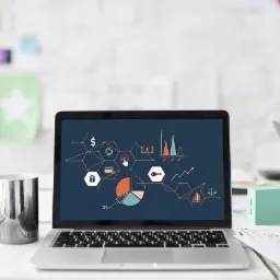 Laptop z wyświetlonymi grafikami analitycznymi, obok kubek, drugi laptop i notatki na biurku w jasnym pomieszczeniu.