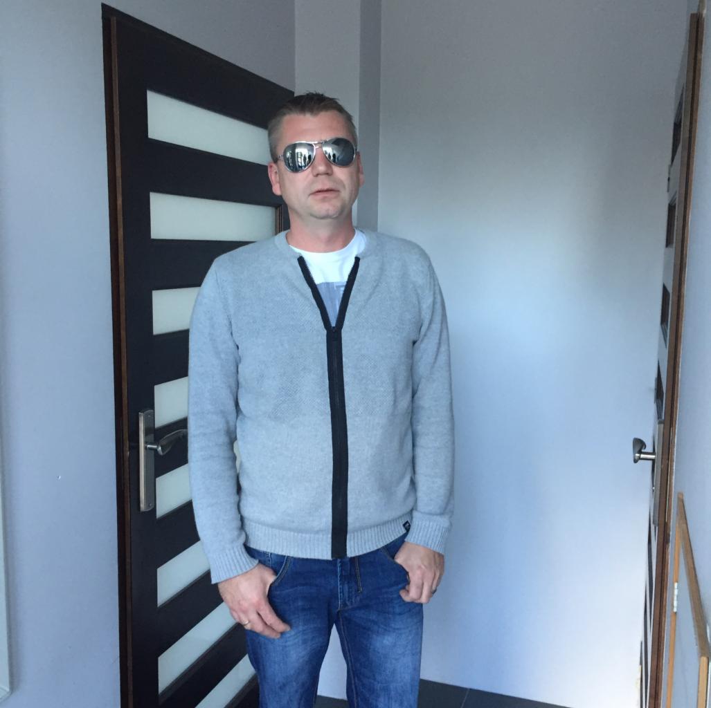 Mężczyzna w okularach przeciwsłonecznych stoi na tle drzwi z poziomymi panelami i jasnej ściany. Ubrany w szary sweter i dżinsy, ręce w kieszeniach.