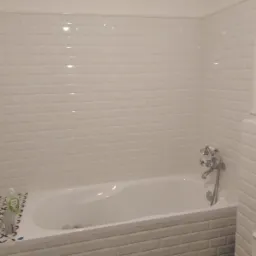 Biała wanna obudowana płytkami typu metro, z chromowaną baterią i prysznicem, otoczona ścianami wyłożonymi tymi samymi płytkami, z widocznym fragmentem toalety i butelką płynu.