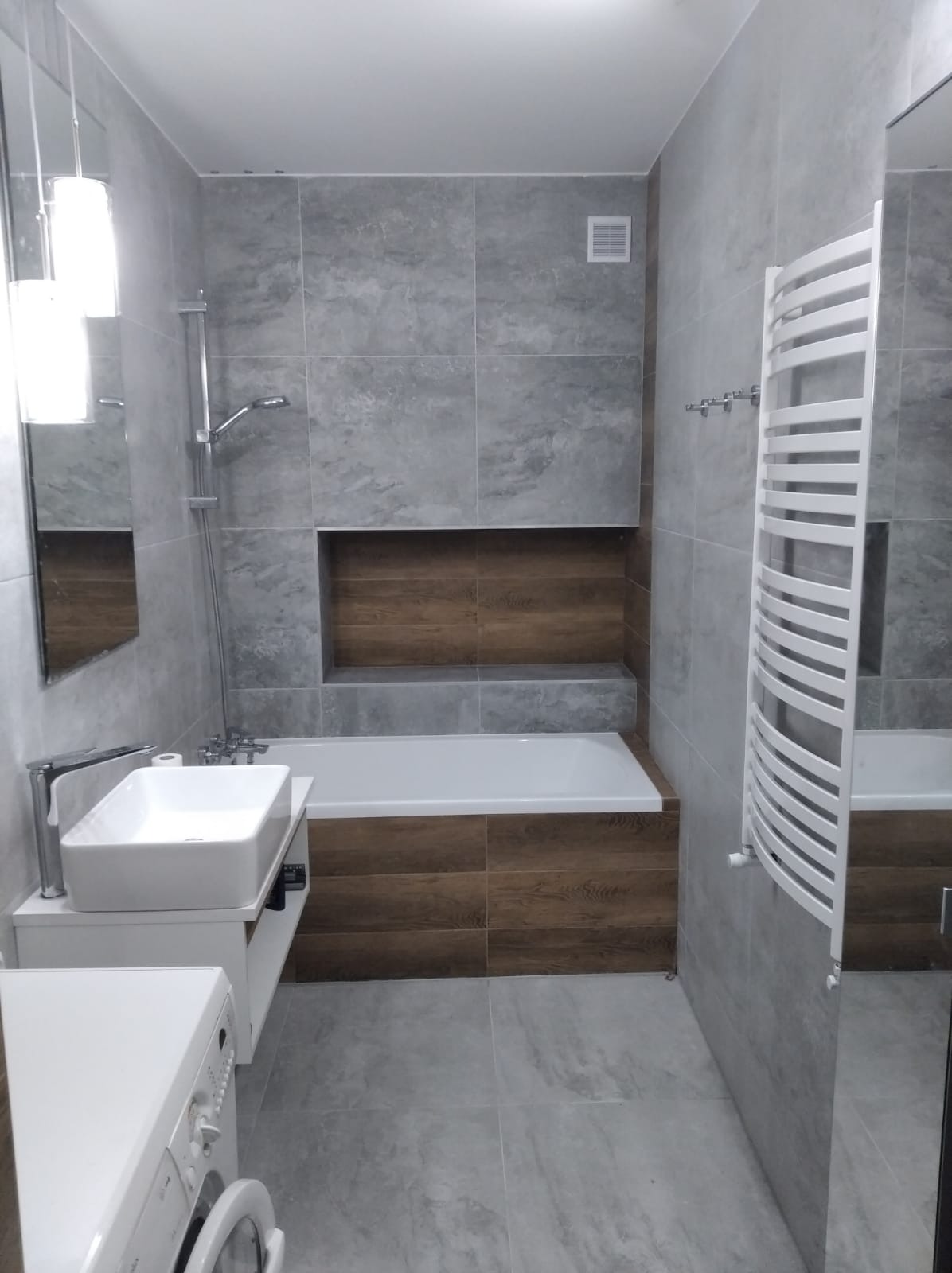 Nowoczesna łazienka z szarymi płytkami imitującymi kamień, zabudową wanny z drewnopodobnych paneli, białą wanną, umywalką nablatową i grzejnikiem drabinkowym.