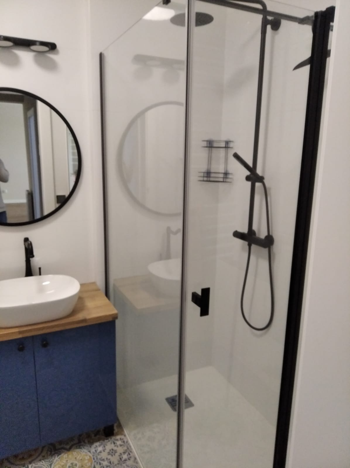 Nowoczesna łazienka z kabiną prysznicową typu walk-in z czarnymi elementami, umywalką nablatową na szafce w kolorze granatowym i okrągłym lustrem.