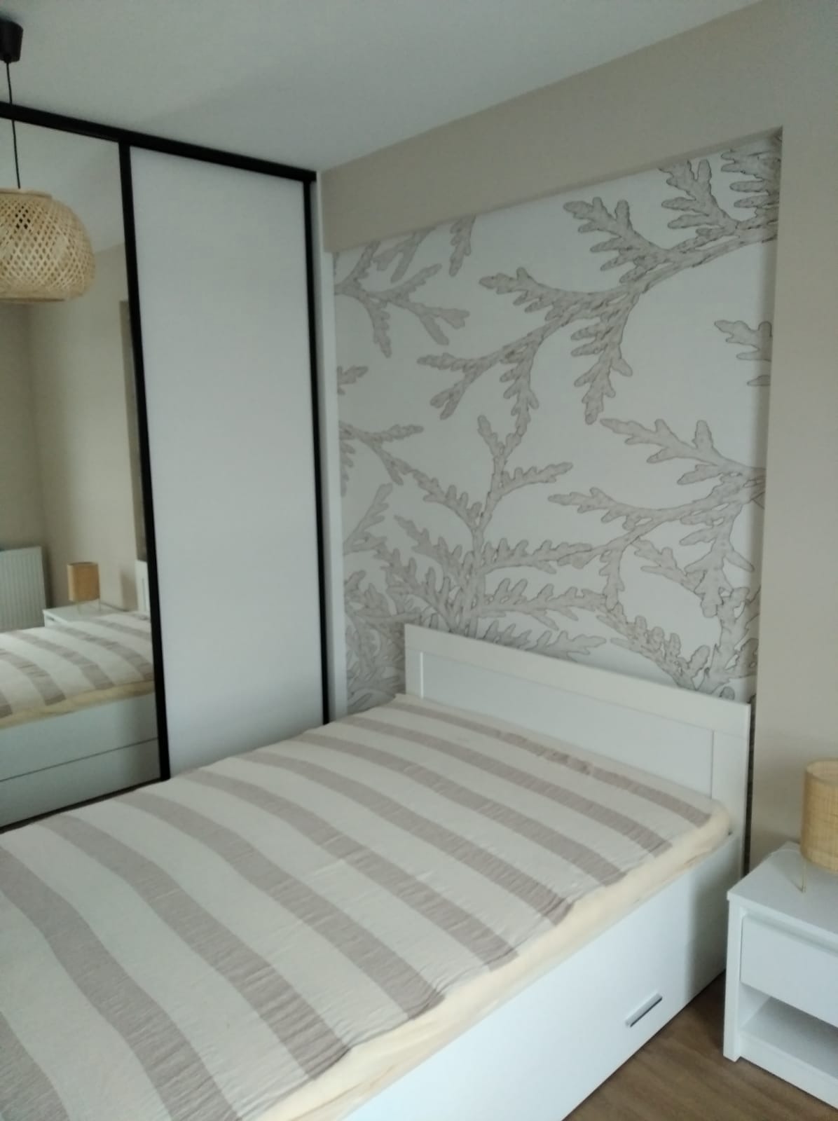 Sypialnia z łóżkiem w paski, szafą z lustrem i ścianą z tapetą w motywy roślinne, oświetlona lampą z naturalnego materiału.