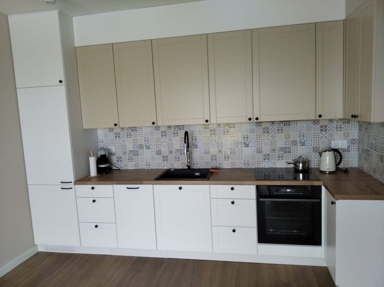 Nowoczesna kuchnia z białymi szafkami dolnymi i beżowymi górnymi, wzorzyste płytki na ścianie, czarny zlew i płyta indukcyjna, drewniany blat.