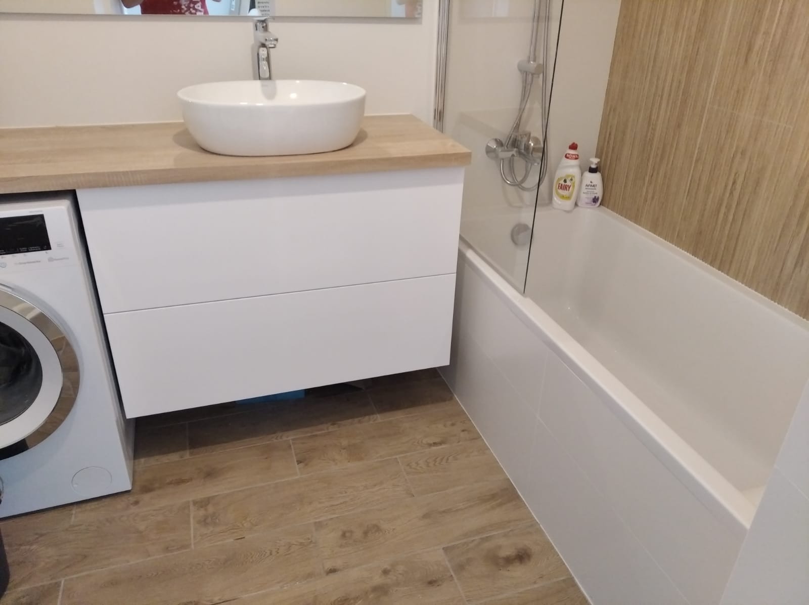 Nowoczesna łazienka po remoncie: umywalka nablatowa na szafce z blatem drewnopodobnym, obok pralka, w tle wanna z parawanem i drewnopodobne płytki na ścianie.